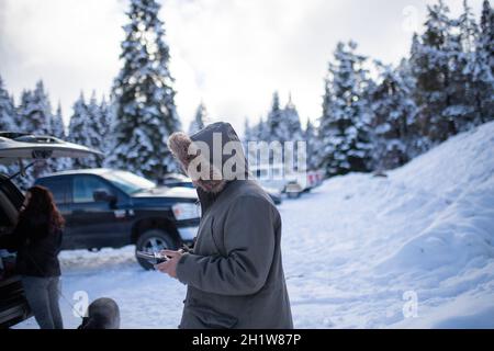 Uomo con cappuccio che tiene il telecomando drone con una bella foresta innevata e camion come sfondo. Uomo misterioso che controlla il drone nei boschi congelati. Adve Foto Stock