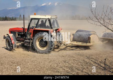 Liptovsky Hradok, Slovacchia - Aprile 22nd, 2018: il trattore svolta durante il traino di pesanti rullo metallico, preparazione di campo secco in primavera, nube di polvere dietro, Foto Stock