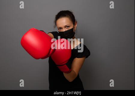 Ritratto di boxer femmina indossando maschera medica nera e guanti rosso boxe, facendo colpo diretto. Pugile donna che puncia verso la macchina fotografica. Concetto di colpo a. Foto Stock