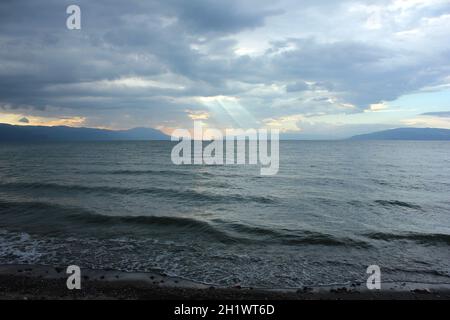 Cielo nuvoloso con sfumature e linee di luce del sole che cadono sulle onde del mare e delle montagne sullo sfondo lontano. Foto Stock