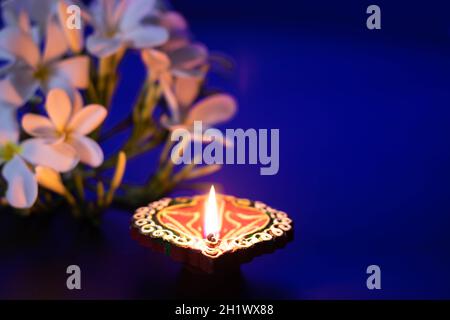 Abito ecologico artigianale Clay Diya Deep dia incandescente con fiori bianchi su sfondo blu. Tema del Festival indiano per Diwali Pooja, Navratri, Dussehra Foto Stock