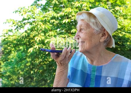 Donna anziana di charme in occhiali rettangolari neri con funzione di riconoscimento vocale sul telefono cellulare. Donna anziana moderna con gad elettronico Foto Stock