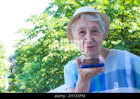 Donna anziana di charme in occhiali rettangolari neri con funzione di riconoscimento vocale sul telefono cellulare. Donna anziana moderna con gad elettronico Foto Stock