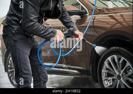 Vista ritagliata dell'uomo che tiene l'attrezzo speciale per lavare l'automobile con acqua ad alta pressione. Le mani del ragazzo puliscono la ruota anteriore di auto con acqua fluente dalla pistola a spruzzo di pressione. Foto Stock