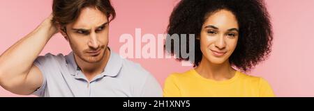 allegra donna latina sorridente alla macchina fotografica vicino confuso uomo che tocca testa isolato su rosa, banner Foto Stock