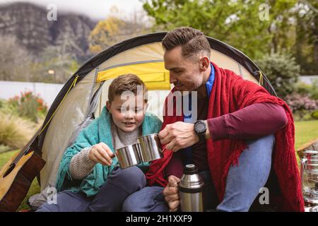 Padre e figlio caucasici tostano le tazze di caffè mentre si siedono in una tenda in giardino Foto Stock