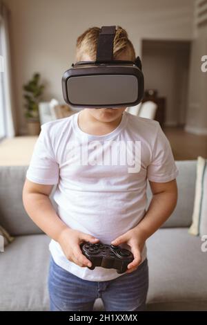 Ragazzo caucasico che indossa un visore vr e tiene il controller di gioco a casa per giocare ai videogiochi Foto Stock