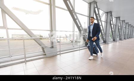 Pronto per il viaggio. Sorridente bell'uomo a piedi medio-orientale con Suitcase in aeroporto Foto Stock