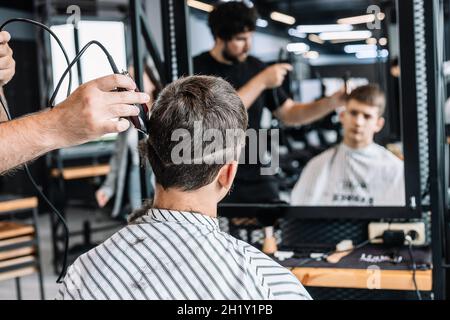 Bello alto barbiere bearded bruna dà un haircut trendy ad un adolescente. Riflessione nello specchio. Taglio professionale e styling per uomo in un barbe Foto Stock