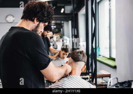 Bello alto barbiere bearded bruna dà un haircut trendy ad un adolescente. Riflessione nello specchio. Taglio professionale e styling per uomo in un barbe Foto Stock