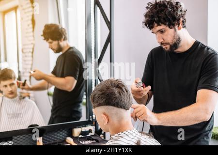 Bello alto barbiere bearded bruna dà un haircut trendy ad un adolescente. Riflessione nello specchio. Taglio professionale e styling per uomo in un barbe Foto Stock