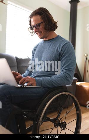 Uomo disabile caucasico che indossa occhiali seduti su sedia a rotelle utilizzando un computer portatile a casa Foto Stock