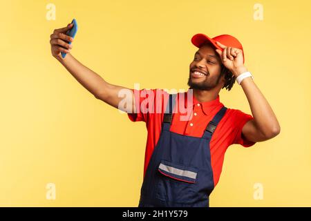 Lavoratore soddisfatto in tute che prende selfie o parla di videochiamata, guardando giocosamente la fotocamera del dispositivo per garantire al cliente la qualità degli ordini online. Studio interno girato isolato su sfondo giallo. Foto Stock