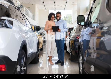 Giovane coppia nera che sceglie l'automobile nel centro di concessionaria moderno, che verifica le caratteristiche del veicolo Foto Stock