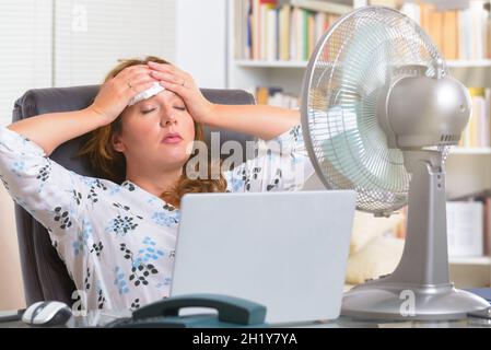 La donna soffre di calore mentre si lavora in ufficio e si tenta di raffreddarsi dalla ventola Foto Stock