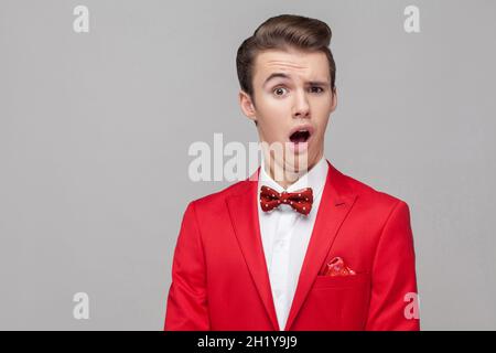 Ritratto di divertente uomo sorpreso con acconciatura elegante in tuxedo rosso e bow tie fissando a sorpresa la fotocamera con comico gimace dumb, facendo un volto. Studio interno girato isolato su sfondo grigio Foto Stock