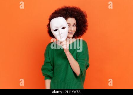 Donna con acconciatura Afro in maglione verde che rimuove la maschera bianca dal volto mostrando la sua espressione sorridente, buon umore, fingendo di essere un'altra persona. Studio interno girato isolato su sfondo arancione. Foto Stock