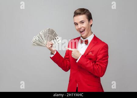 Guarda i miei soldi. Ritratto di uomo estremamente felice con capelli alla moda in giacca rossa e arco cravatta puntando a un mazzo di dollari, esprimendo gioia, eccitazione. studio girato isolato su sfondo grigio Foto Stock