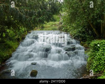 Una piccola cascata a più livelli circondata da alberi in un parco suburbano, presa con un otturatore lento per catturare la sussurrazione dell'acqua che cade. Foto Stock