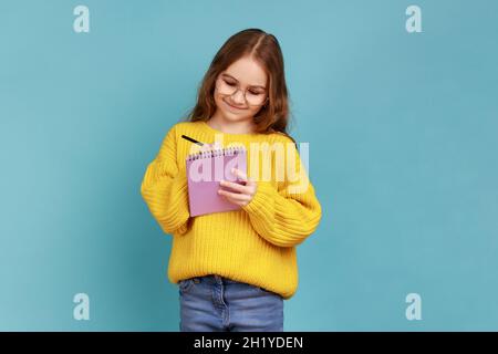 Ritratto di positiva piccola ragazza che scrive in taccuino, sorridente, note, bambino di scuola che fa i compiti, indossando il maglione giallo di stile casual. Studio interno girato isolato su sfondo blu. Foto Stock