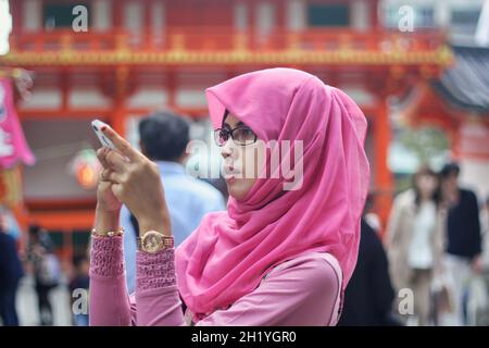 Primo piano ritratto di giovane musulmana asiatica che indossa hijab rosa e occhiali tenendo e guardando smartphone con sfondo bokeh in strada. Foto Stock