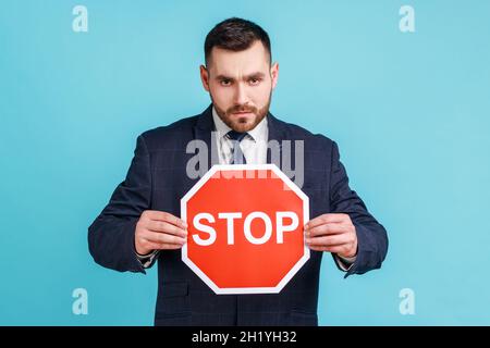Uomo serio preoccupato che indossa abiti di stile ufficiale tenendo rosso stop segno in mani, attenzione, attenzione concetto attento, avvertenza, guardando la fotocamera. Studio interno girato isolato su sfondo blu. Foto Stock