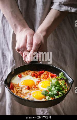 Piatti della cucina tradizionale israeliana Shakshuka (uova fritte con verdure, pomodori e paprika in padella di ghisa in mani femminili) Foto Stock