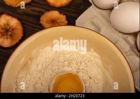 Un uovo di pollo bianco rotto in una ciotola con farina di frumento, patatine secche di mela e diverse uova intere in un vassoio di cartone bianco, primo piano selettivo Foto Stock