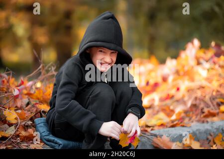 sly boy in una giacca nera con cappuccio siede sul marciapiede e sorride Foto Stock