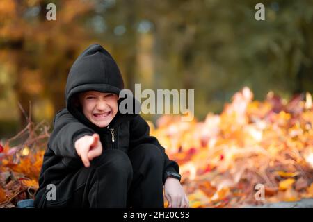 sly boy in una giacca nera con cappuccio siede sul marciapiede e sorride Foto Stock