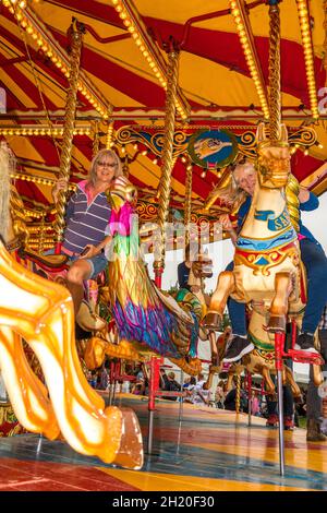 Gli amici che si divertono su una giostra tradizionale cavallo giostra andare rotondo. I gallo di vapore si cavalcano con i cavalli in un giro sul fairground Foto Stock