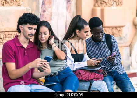 Quattro giovani studenti diversi che usano il cellulare mentre si siedono all'aperto Foto Stock