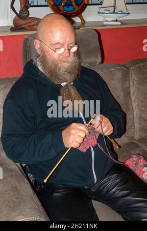 Mann mit Langem Bart und Brille sitzt auf der Couch und strickt einen Schal Foto Stock