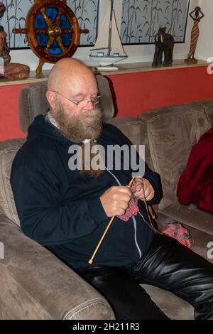 Mann mit Langem Bart und Brille sitzt auf der Couch und strickt einen Schal Foto Stock