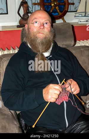 Mann mit Langem Bart und Brille sitzt auf der Couch und strickt einen Schal Foto Stock