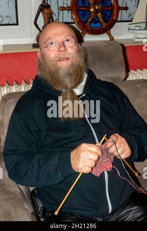 Mann mit Langem Bart und Brille sitzt auf der Couch und strickt einen Schal Foto Stock