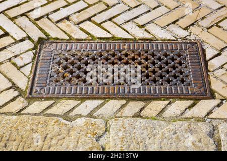 ferro arrugginito manhole rettangolare con fori sagomati in forma di croci nel coperchio sul marciapiede pavimentato con mattonelle di mattoni, vecchio portello della fogna su un marciapiede di pietra Foto Stock