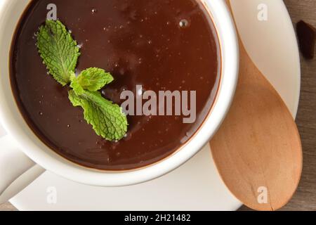 Macrofumo di cioccolato caldo in tazza di ceramica bianca su un piatto con cucchiaio di legno e foglie di menta su tavola di legno. Vista dall'alto. Composizione orizzontale. Foto Stock