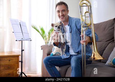 L'uomo con il suo cane mentre pratica il trombone nel suo tempo libero nel salotto di casa. Vista frontale. Composizione orizzontale. Foto Stock