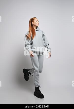 Donna allegra e sottile con capelli rossi con acconciatura a coda di cavallo in felpa con cappuccio, pantaloni e scarpe nere alla moda, corse Foto Stock