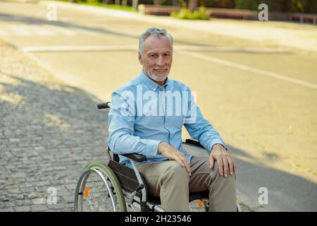 Uomo anziano in sedia a rotelle sorridente a macchina fotografica mentre cavalca da solo nel parco in una giornata estiva Foto Stock