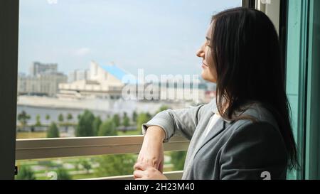 la giovane donna in un abito da lavoro si alza da una finestra aperta e guarda in lontananza Foto Stock
