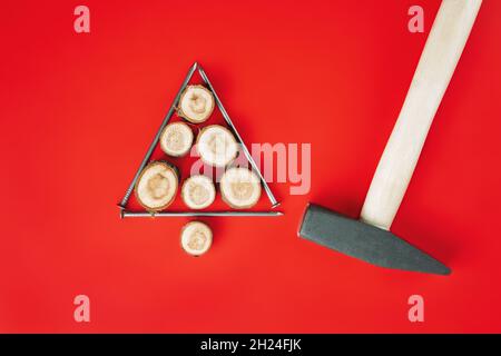 Chiodi lunghi e tagli di legno o lastre di ramo, disposti a forma di albero di Natale con martello su sfondo rosso. Disposizione piatta Foto Stock