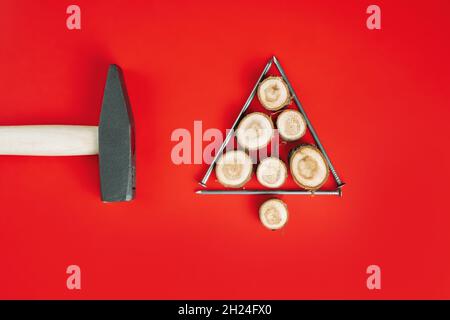 Chiodi lunghi e tagli di legno o lastre di ramo, disposti a forma di albero di Natale con martello su sfondo rosso. Disposizione piatta Foto Stock