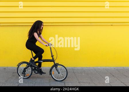 Giovane donna nera con capelli lunghi e vestito nero che guida la sua bicicletta di fronte A Un muro giallo Foto Stock