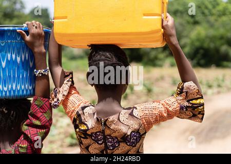 Vista posteriore di una giovane ragazza africana nera in un vestito colouful con un'acqua enorme può sulla sua testa che cammina su strada sterrata Foto Stock