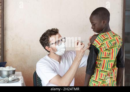 Medico con maschera protettiva per il viso preparazione del sito di iniezione sul braccio di un coraggioso scolaro africano nero in oder per somministrare un f Foto Stock