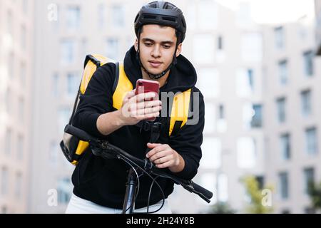 Corriere maschio appoggiato sul manubrio di una bicicletta indossando zaino per la consegna di cibo e casco da bicicletta guardando il telefono cellulare Foto Stock