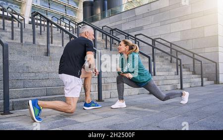 Scatto a tutta lunghezza di coppia sportiva di mezza età, uomo e donna che allunga le gambe mentre si esercitano insieme all'aperto in una giornata di sole Foto Stock