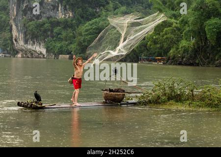 Un pescatore cormorano in un cappello conico su una zattera di bambù lancia una rete di getto nel fiume li, Xingping, Cina. Foto Stock